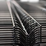 Nærbillede af en række Panelhegn 3D 5/5mm - HF PREMIUM Pulverlakeret Sort RAL 9005 trådstativer stablet sammen. Fokus er på de indviklede, gentagne linjer dannet af ledningerne, hvilket giver en følelse af dybde og strukturelt mønster. Baggrunden er sløret, hvilket fremhæver detaljerne i trådkonstruktionen.