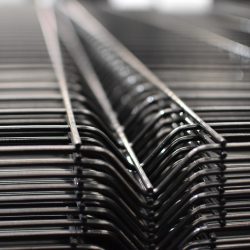 Nærbillede af en række Panelhegn 3D 5/5mm - HF PREMIUM Pulverlakeret Sort RAL 9005 trådstativer stablet sammen. Fokus er på de indviklede, gentagne linjer dannet af ledningerne, hvilket giver en følelse af dybde og strukturelt mønster. Baggrunden er sløret, hvilket fremhæver detaljerne i trådkonstruktionen.