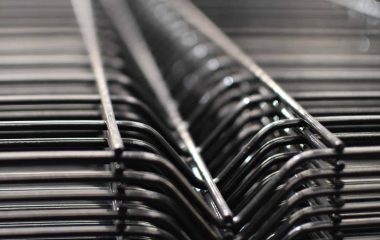 Nærbillede af en række Panelhegn 3D 5/5mm - HF PREMIUM Pulverlakeret Sort RAL 9005 trådstativer stablet sammen. Fokus er på de indviklede, gentagne linjer dannet af ledningerne, hvilket giver en følelse af dybde og strukturelt mønster. Baggrunden er sløret, hvilket fremhæver detaljerne i trådkonstruktionen.