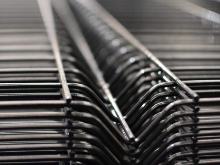 Nærbillede af en række Panelhegn 3D 5/5mm - HF PREMIUM Pulverlakeret Sort RAL 9005 trådstativer stablet sammen. Fokus er på de indviklede, gentagne linjer dannet af ledningerne, hvilket giver en følelse af dybde og strukturelt mønster. Baggrunden er sløret, hvilket fremhæver detaljerne i trådkonstruktionen.