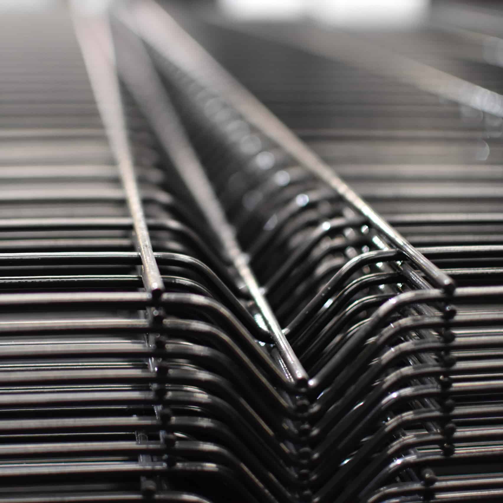 Nærbillede af en række Panelhegn 3D 5/5mm - HF PREMIUM Pulverlakeret Sort RAL 9005 trådstativer stablet sammen. Fokus er på de indviklede, gentagne linjer dannet af ledningerne, hvilket giver en følelse af dybde og strukturelt mønster. Baggrunden er sløret, hvilket fremhæver detaljerne i trådkonstruktionen.