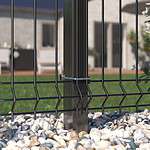 Nærbillede af Metal Hegn 3D Panel "HF-BUDGET SERIES" i en pulverlakeret sort finish, med lodrette stålstolper, fastgjort med metalbeslag. Hegnet er omgivet af små hvide og grå småsten. I baggrunden er der en sløret udsigt til et hus og en grøn græsplæne.