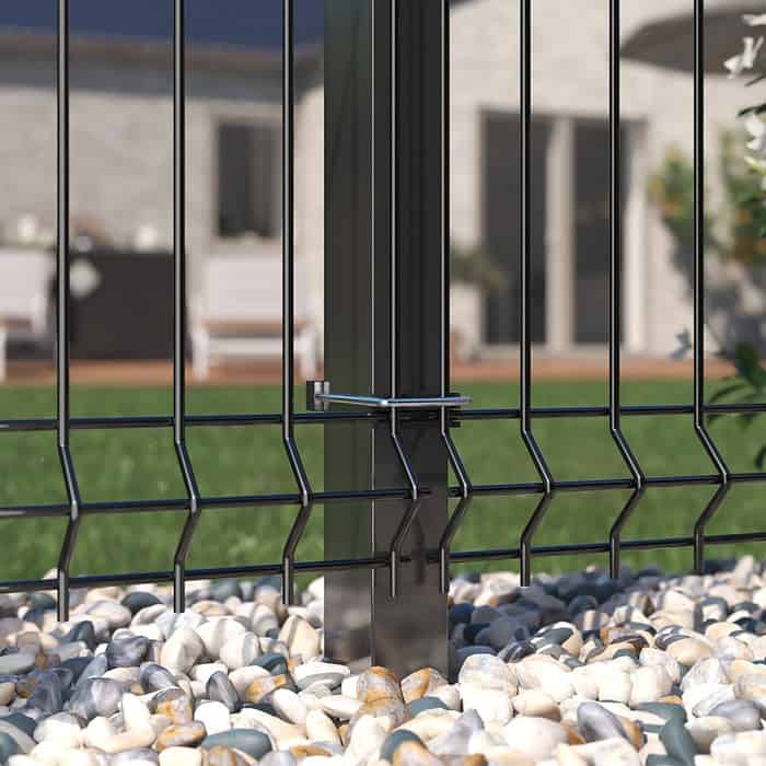 Nærbillede af Metal Hegn 3D Panel "HF-BUDGET SERIES" i en pulverlakeret sort finish, med lodrette stålstolper, fastgjort med metalbeslag. Hegnet er omgivet af små hvide og grå småsten. I baggrunden er der en sløret udsigt til et hus og en grøn græsplæne.