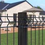 Nærbillede af et Metal Hegn 3D Panel "HF-BUDGET SERIES" Inkl. Stålstolper & Beslag - Pulverlakeret Sorter hegnspæl og trådnet hegn foran hus med terrasse. Det hvide hus har store vinduer, mens baggrunden afslører havemøbler og et delvist skyggefuldt område, hvilket tyder på en solskinsdag.