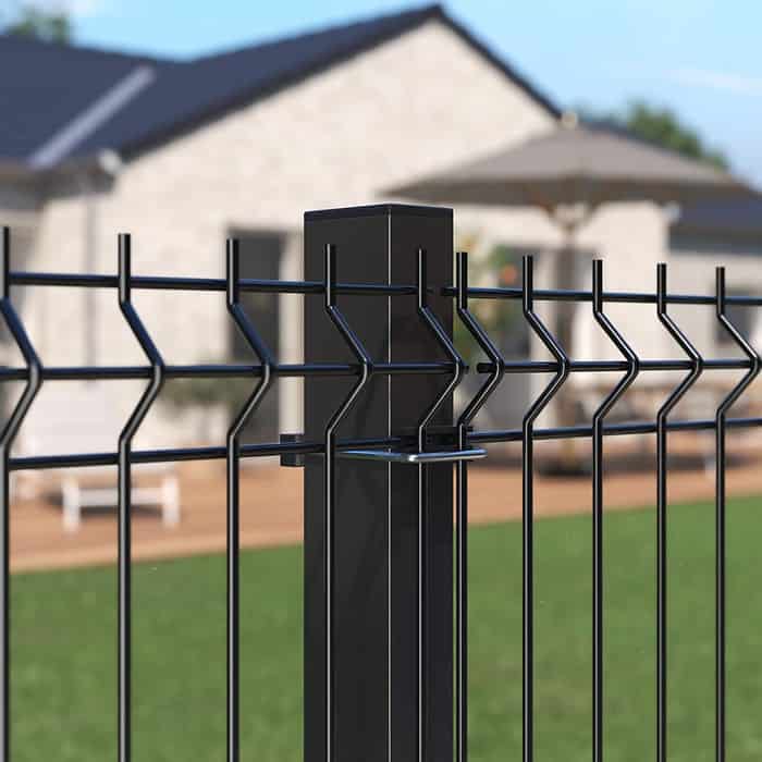 Nærbillede af et Metal Hegn 3D Panel "HF-BUDGET SERIES" Inkl. Stålstolper & Beslag - Pulverlakeret Sorter hegnspæl og trådnet hegn foran hus med terrasse. Det hvide hus har store vinduer, mens baggrunden afslører havemøbler og et delvist skyggefuldt område, hvilket tyder på en solskinsdag.