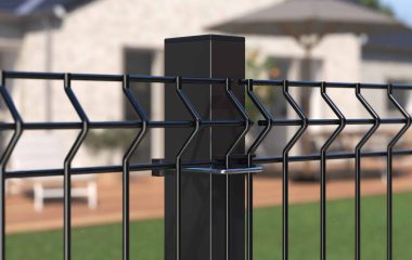 Detaljeret visning af et Metal Hegn 3D Panel "HF-BUDGET SERIES" Inkl. Stålstolper & Fittings i Pulverlakeret Sort, med et geometrisk design, med en velholdt græsplæne i forgrunden og et moderne et-plans hus med et stort dæk og udendørsmøbler i baggrunden. Den klare himmel indikerer, at det er en solskinsdag.