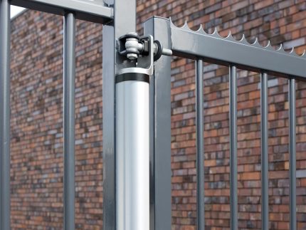 Nærbillede af et metallisk porthængsel med en hydraulisk lukker fastgjort til et gråt jernhegn. 2D Panellåge "TREND" Galvaniseret/Pulverlakeret har lodrette stænger og spidse toppe. I baggrunden er der en murstensvæg med et brunt og rødt mønster. Billedet vises i dagslys.