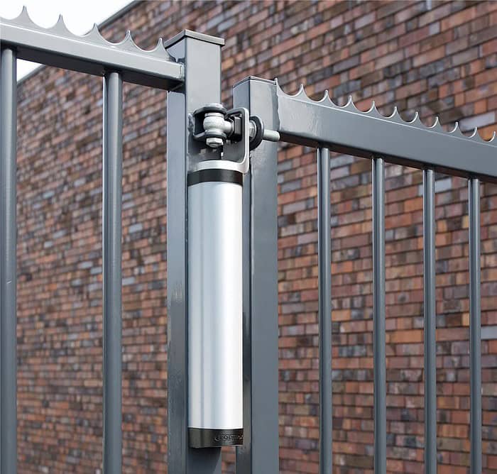Nærbillede af et metallisk porthængsel med en hydraulisk lukker fastgjort til et gråt jernhegn. 2D Panellåge "TREND" Galvaniseret/Pulverlakeret har lodrette stænger og spidse toppe. I baggrunden er der en murstensvæg med et brunt og rødt mønster. Billedet vises i dagslys.