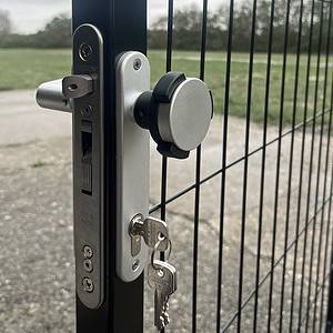 Nærbillede af en metallås på et sort hegn med en sølv KIDLOC - Børnesikret Låsehåndtag til Institutioner og to nøgler indsat. Baggrunden viser en asfalteret sti og en græsmark med nogle træer i det fjerne. Himlen ser overskyet ud.