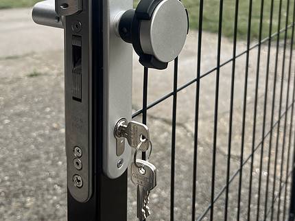 Nærbillede af en metallås på et sort hegn med en sølv KIDLOC - Børnesikret Låsehåndtag til Institutioner og to nøgler indsat. Baggrunden viser en asfalteret sti og en græsmark med nogle træer i det fjerne. Himlen ser overskyet ud.