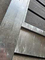 Nærbillede af en metal- og træstruktur, der viser hjørnet, hvor en galvaniseret metallisk bjælke skærer træpaneler. Overfladerne udviser en vis tekstur og slid, hvor metalliske dele, herunder Bundplade Galvaniseret Stål - 1815x175x35/2mm, viser en smule misfarvning og ridser. Billedvinklen er lidt skæv.