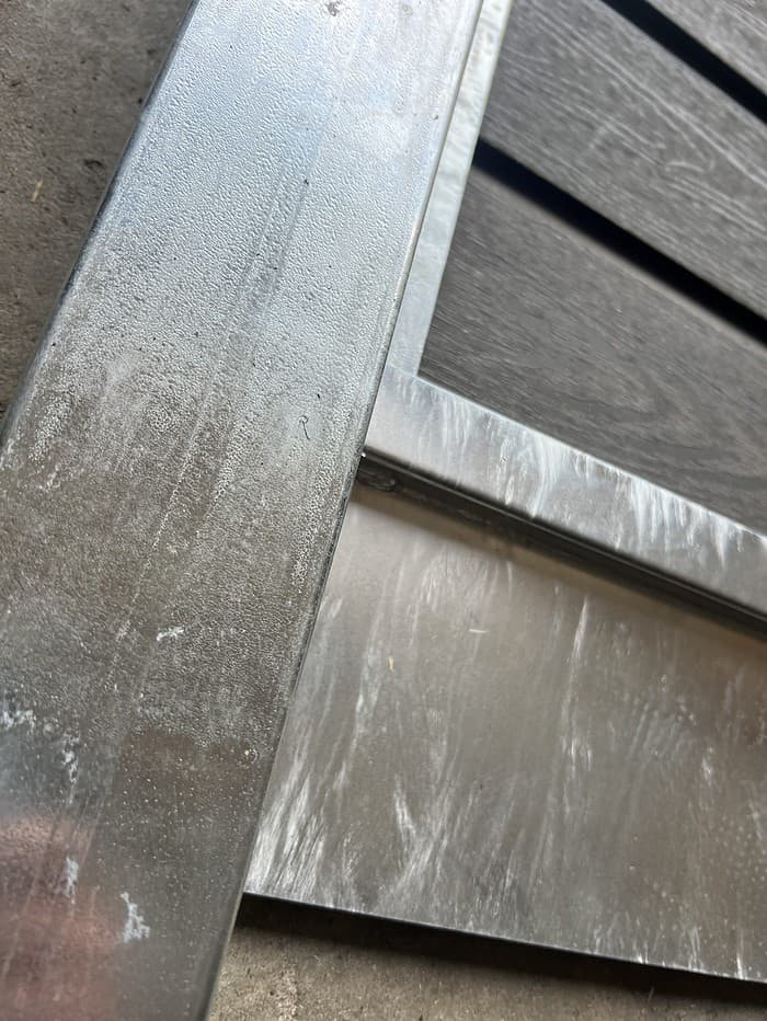 Nærbillede af en metal- og træstruktur, der viser hjørnet, hvor en galvaniseret metallisk bjælke skærer træpaneler. Overfladerne udviser en vis tekstur og slid, hvor metalliske dele, herunder Bundplade Galvaniseret Stål - 1815x175x35/2mm, viser en smule misfarvning og ridser. Billedvinklen er lidt skæv.