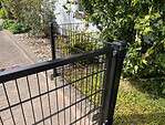 Et sort Panelhegn 2D STANDARD 6/5/6 mm metalhegn, udformet som et panel med en åben port, står ved siden af et fortov og grønne buske, der fører hen mod en bygning med hvide vægge. Her og der ses lilla blomster blandt det grønne.