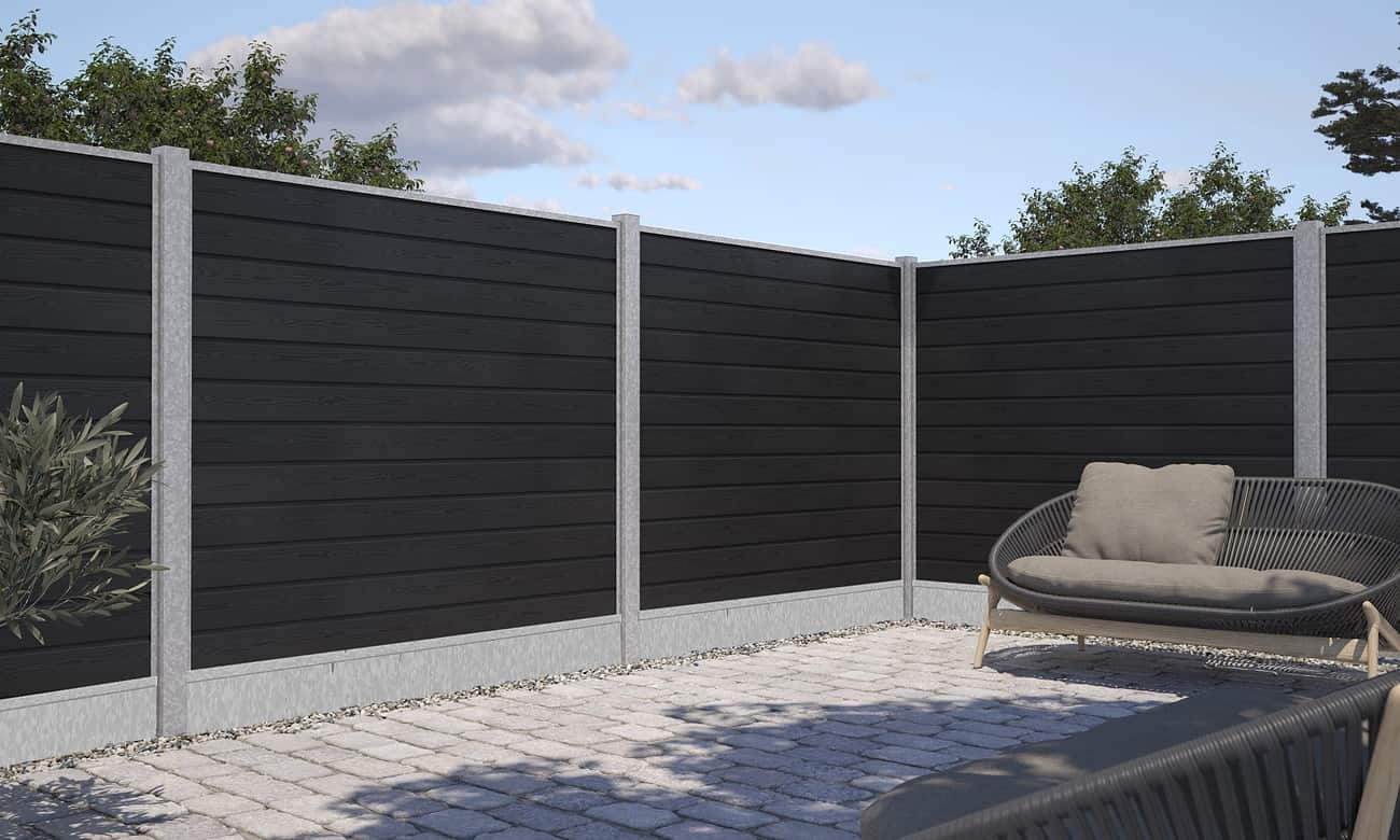 Et minimalistisk udendørs hjørnerum har et højt, sort træhegn med grå betonpæle og grus i bunden. En kurvestol med beige hynder hviler på en stenbelagt terrasse, komplet med Bundplade Galvaniseret Stål - 1815x175x35/2mm accenter. Grønt er synligt i baggrunden under en delvist overskyet himmel.