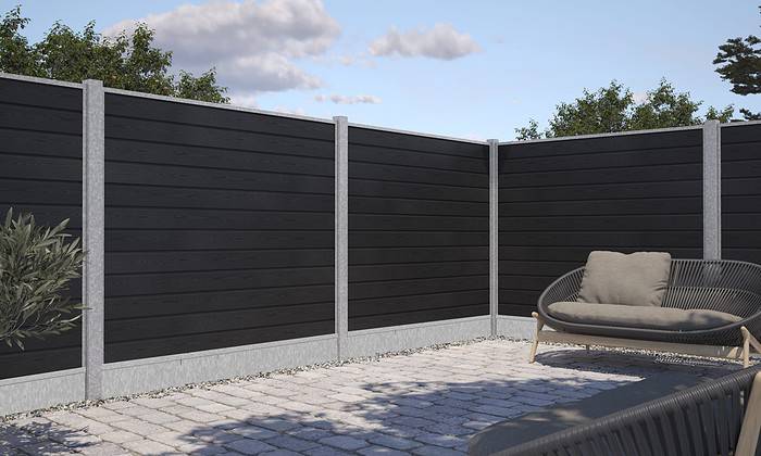 Et minimalistisk udendørs hjørnerum har et højt, sort træhegn med grå betonpæle og grus i bunden. En kurvestol med beige hynder hviler på en stenbelagt terrasse, komplet med Bundplade Galvaniseret Stål - 1815x175x35/2mm accenter. Grønt er synligt i baggrunden under en delvist overskyet himmel.