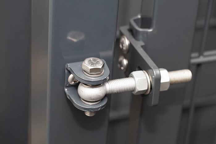 Nærbillede af en hængselmekanisme i metal, monteret på en grå Dobbeltlåge »TREND«, komplet med Locinox-låsekasse, herunder bolte, møtrikker og en cylindrisk stang, der sikrer stabil fastgørelse og bevægelse.
