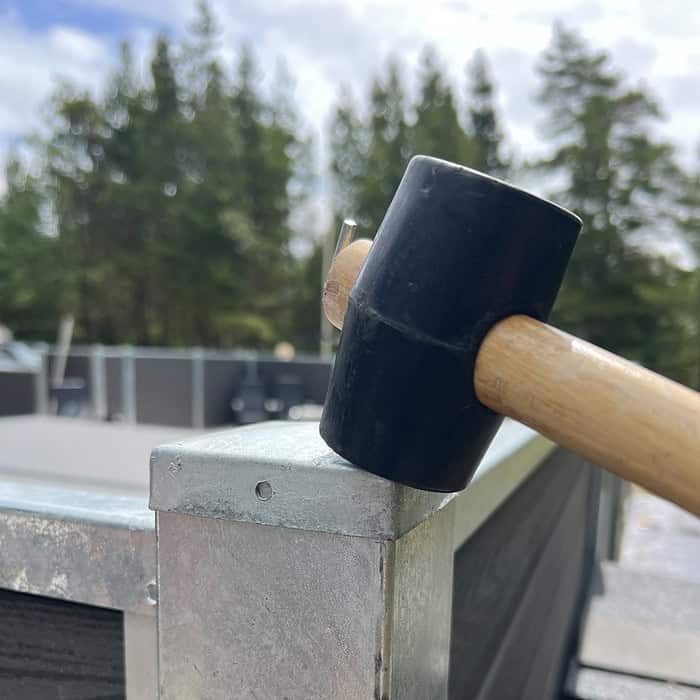 En sort gummihammer med træhåndtag banker på et Stolpehat Varm galvaniseret stål HDG 80x80mm hjørne, med en skov og nogle byggematerialer i den slørede baggrund. Billedet fokuserer på værktøjet og metalkanten og fremhæver byggeaktiviteten.