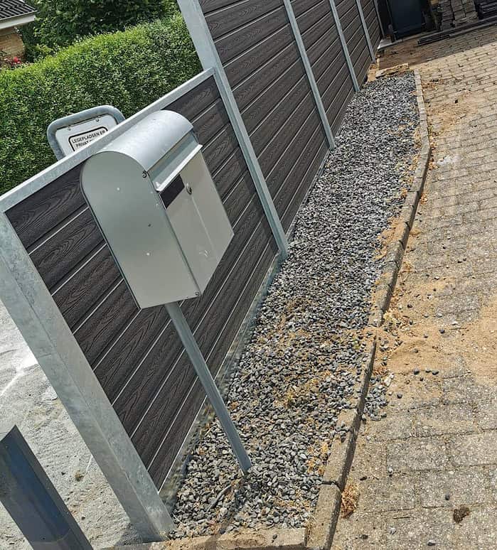 En høj sølvpostkasse med Bundplade Galvaniseret Stål - 1815x175x35/2mm er monteret på en stang ved siden af en gangsti, som har gråt grus på den ene side og beige sand på den anden. Postkassen står foran et mørkt hegn, med grønne buske synlige i baggrunden.