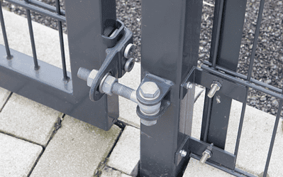 Nærbillede af et sort metal porthængsel og låsesystem med Dobbeltlåge "TREND" Komplet med Locinox låsekasse. Porten er sikret med bolte og skruer og forbundet til et sort metalhegn. Jorden er brolagt med betonblokke, der komplementerer TREND-designet.