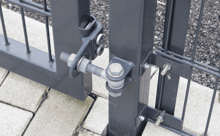 Nærbillede af et sort metal porthængsel og låsesystem med Dobbeltlåge "TREND" Komplet med Locinox låsekasse. Porten er sikret med bolte og skruer og forbundet til et sort metalhegn. Jorden er brolagt med betonblokke, der komplementerer TREND-designet.