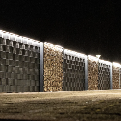 En hvid bil er parkeret ved siden af en lang, veloplyst væg lavet af stablede brændesektioner og store panelhegn. Billedet, der er taget om natten, har "LightUp" LED-Lysliste, der oplyser væggen og kaster skygger på jorden.