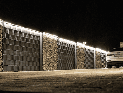 En hvid bil er parkeret ved siden af en lang, veloplyst væg lavet af stablede brændesektioner og store panelhegn. Billedet, der er taget om natten, har "LightUp" LED-Lysliste, der oplyser væggen og kaster skygger på jorden.