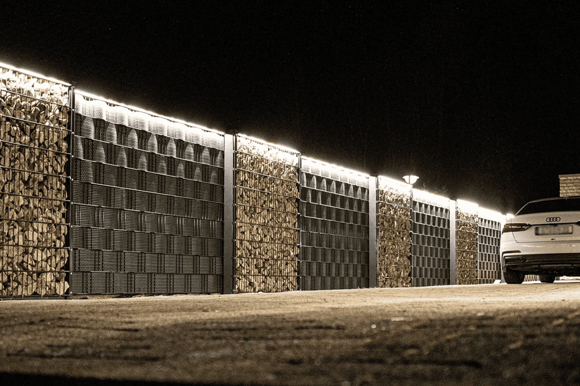 En hvid bil er parkeret ved siden af en lang, veloplyst væg lavet af stablede brændesektioner og store panelhegn. Billedet, der er taget om natten, har "LightUp" LED-Lysliste, der oplyser væggen og kaster skygger på jorden.