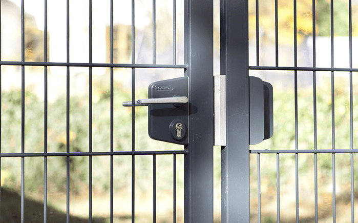 Et nærbillede af den moderne sorte Dobbeltlåge "TREND"-port, sammensat af slanke lodrette og vandrette metalstænger med en sikker Locinox låsekasse-mekanisme. Baggrunden er let sløret og viser grønt og et hegn - et perfekt eksempel på moderne design.