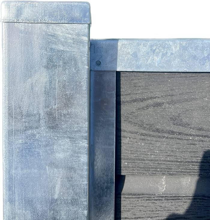 Et nærbillede af en metallisk hjørnestruktur, med en lodret metalstolpe til venstre og en vandret bjælke på toppen, begge i galvaniseret stål med en forvitret sølvskinnende finish. Stolpehat Varm galvaniseret stål HDG 80x80 mm passer pænt oven på 80x80 mm stolpen og indrammer et mørkt tekstureret panel på højre side af billedet.