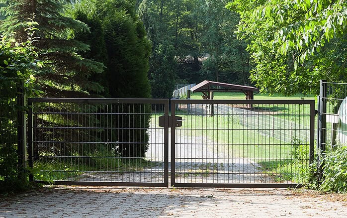 En Dobbeltlåge "TREND" komplet med Locinox låsekasse blokerer indgangen til en indkørsel, der fører til et græsareal med et træskur, alt sammen omgivet af træer og grønne områder på en solrig dag.