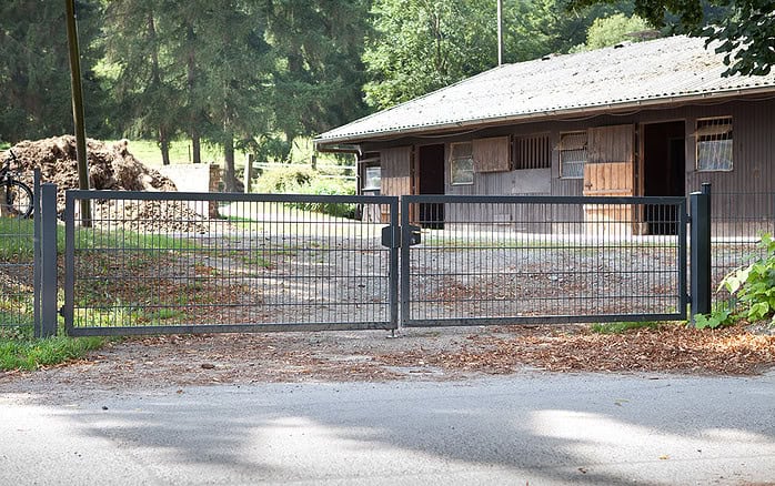 Dobbeltlåge "TREND" Komplet med Locinox låsekasse spærrer en grusindkørsel foran en brun staldbygning med åbne vinduer, omgivet af træer og grønne områder.