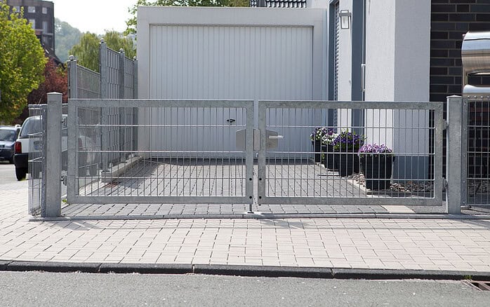 En Dobbeltlåge "TREND" Komplet med Locinox låsekasse blokerer indkørslen til en lukket hvid garage. Fire lilla blomster i potter står i højre side ved siden af en moderne, hvid bygning.