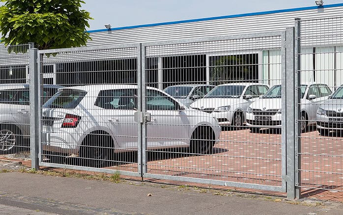 Flere hvide biler er parkeret på en indhegnet plads uden for en moderne industribygning, og en Dobbeltlåge "TREND" komplet med Locinox låsekasse er synlig nær fortovet og træet i forgrunden.