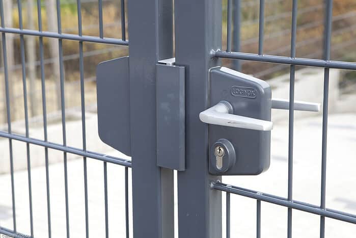 Et nærbillede af den grå metal-dobbeltlåge »TREND«, komplet med Locinox-låsekasse, med lodrette og vandrette stænger, opstillet udendørs tæt på et brolagt og indhegnet område.