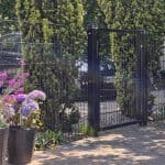 En Metallåge 2D Panelfyld 6/5/6mm Sort Ral 9005 Komplet med netdesign står mellem høje grønne buske, med en parkeret bil synlig bag lågen og potteblomster placeret på det asfalterede område foran.