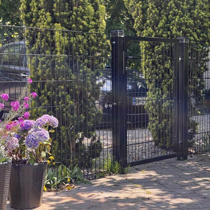 En Metallåge 2D Panelfyld 6/5/6mm Sort Ral 9005 Komplet med netdesign står mellem høje grønne buske, med en parkeret bil synlig bag lågen og potteblomster placeret på det asfalterede område foran.