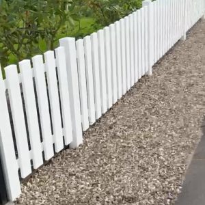 Et hvidt stakit løber langs en grussti med grønne buske og græs på den anden side af hegnet.