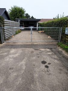 En metalport blokerer indgangen til en indkørsel, der fører til en carport med to biler parkeret nedenunder. Indkørslen er belagt med mursten og flankeret af hække og hegn.