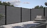 Et moderne udendørs terrasseområde med et højt, gråt privathegn indrammet af Komposit Hegn med sorte pulverlakeret stålstolper & u-skinner - Type 1. Jorden er brolagt med firkantede stenfliser, og der er en sort udendørs sofa med beige hynder til højre. . En lille grøn busk er til venstre, og træer er synlige i baggrunden.