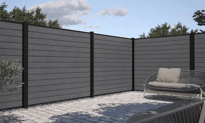 Et moderne udendørs terrasseområde med et højt, gråt privathegn indrammet af Komposit Hegn med sorte pulverlakeret stålstolper & u-skinner - Type 1. Jorden er brolagt med firkantede stenfliser, og der er en sort udendørs sofa med beige hynder til højre. . En lille grøn busk er til venstre, og træer er synlige i baggrunden.