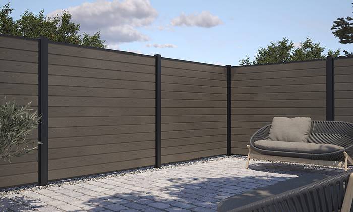 Et moderne indhegnet terrasseområde med vandrette trælameller og sorte metalstolper. Grunden er belagt med lysegrå stenfliser. Der er en hyggelig udendørs stol med hynder i højre side, indrammet af en lækker Komposit Hegn med sorte pulverlakeret stålstolper & u-skinner - Type 1. Træer og en overskyet himmel ses i baggrunden.