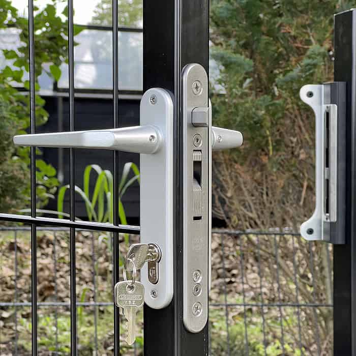 En detaljeret visning af et sølv dørhåndtag og låsemekanisme på en Metallåge 2D Panelfyld 6/5/6mm Sort Ral 9005 Komplet sort metal låge. Den godt sikrede lås har en isat nøgle, og lågen udgør en del af et udendørs indhegnet område med frodigt grønt i baggrunden.