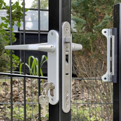 En detaljeret visning af et sølv dørhåndtag og låsemekanisme på en Metallåge 2D Panelfyld 6/5/6mm Sort Ral 9005 Komplet sort metal låge. Den godt sikrede lås har en isat nøgle, og lågen udgør en del af et udendørs indhegnet område med frodigt grønt i baggrunden.