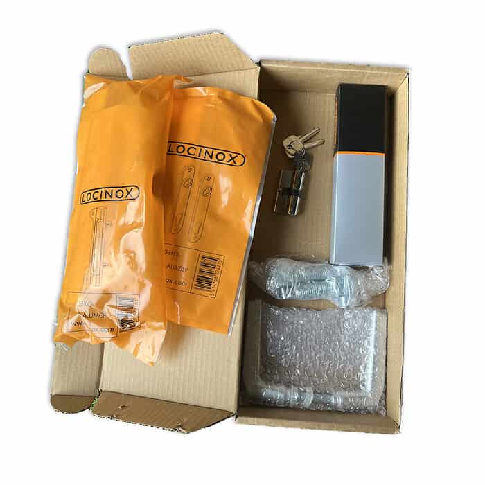 En papkasse indeholdende flere genstande, inklusive to orange Locinox-pakker, et bundt nøgler fastgjort til en cylindrisk metalgenstand, der sandsynligvis er en del af en Metallåge 2D Panelfyld 6/5/6mm Sort Ral 9005 Komplet, en grå rektangulær æske, en boble-omviklet komponent , og et lille bundt skruer i plastfolie.