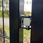 Nærbillede af et sekskantet bolthængsel fastgjort til den sorte ramme, Sort Ral 9005, af Metallåge 2D Panelfyld 6/5/6mm Sort Ral 9005 Komplet med et skovområde i baggrunden. Hængslet forbinder to sektioner af strukturen lavet af metalstænger.