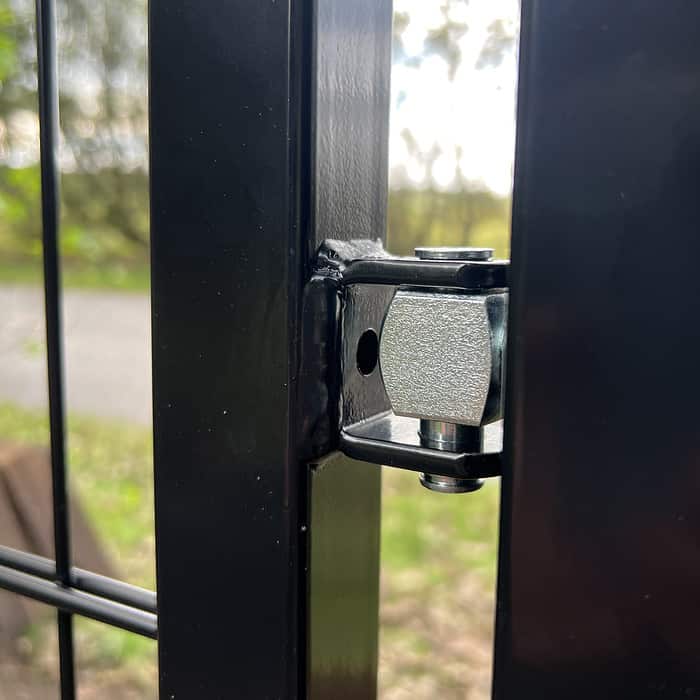 Nærbillede af et sekskantet bolthængsel fastgjort til den sorte ramme, Sort Ral 9005, af Metallåge 2D Panelfyld 6/5/6mm Sort Ral 9005 Komplet med et skovområde i baggrunden. Hængslet forbinder to sektioner af strukturen lavet af metalstænger.