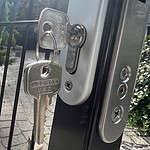 Nærbillede af et sæt nøgler indsat i en udendørs lås på en Metallåge 2D Panelfyld 6/5/6mm færdig i Sort Ral 9005. Nøglerne, indgraveret med "LOCINOX V.S.Z", dingler fra låsen. Porten ser ud til at være placeret i en anlagt have med buske synlige i baggrunden.