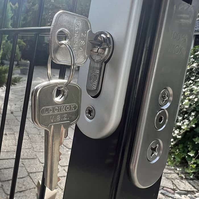 Nærbillede af et sæt nøgler indsat i en udendørs lås på en Metallåge 2D Panelfyld 6/5/6mm færdig i Sort Ral 9005. Nøglerne, indgraveret med "LOCINOX V.S.Z", dingler fra låsen. Porten ser ud til at være placeret i en anlagt have med buske synlige i baggrunden.