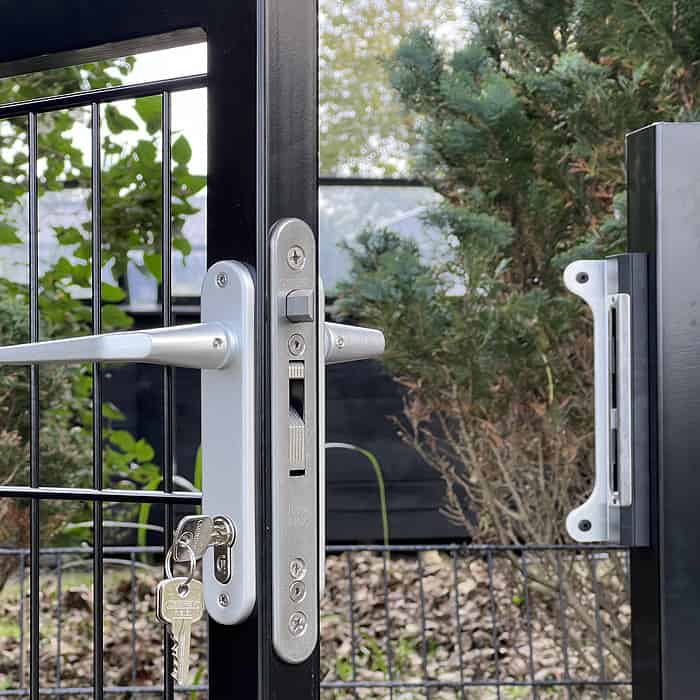 Nærbillede af en metalportlås med en nøgle indsat i låsen og en grøn have i baggrunden. Låsen er sølv og fastgjort til en Metallåge 2D Panelfyld 6/5/6mm Sort Ral 9005 Komplet med trådnet. Der er træer og buske i nærheden, hvilket forstærker den fredfyldte scene.