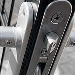 Nærbillede af en sølv dørlåsemekanisme med synlige skruer og en LOBP (Lift Off Bolt) på siden, monteret på en Metallåge 2D Panelfyld 6/5/6mm Sort Ral 9005 Komplet. Låsen er aktiveret og viser bolten udstrakt. Billedet har et gråtonefilter, der fremhæver de metalliske komponenter mod en let sløret baggrund.